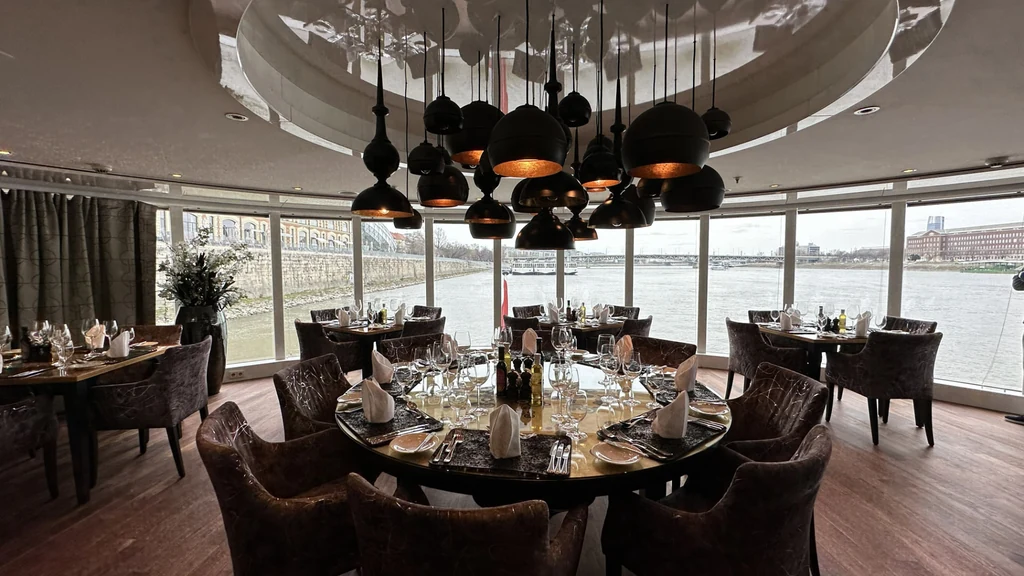 Round dining table set for dinner in a river cruise ship restaurant with panoramic river views.