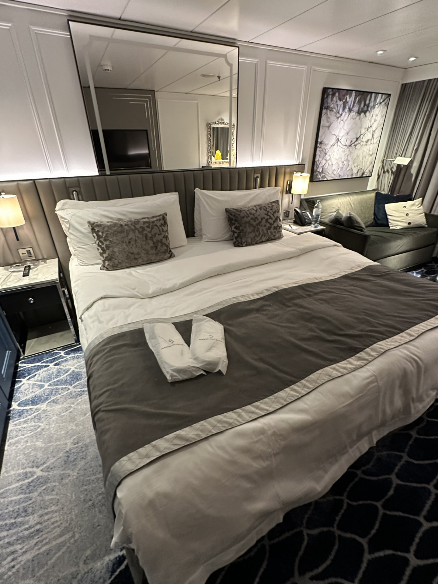 Cruise ship stateroom with a made-up king bed, upholstered headboard, sitting area, and reading lamps