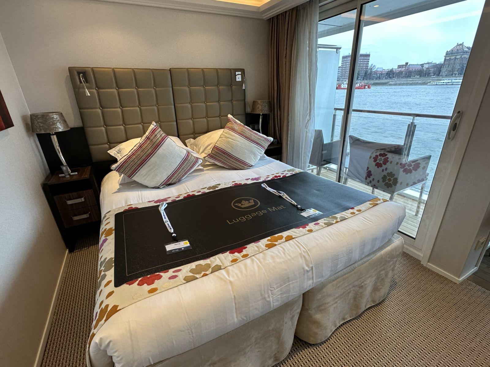 River cruise cabin with a double bed, a luggage mat, and a sliding glass door opening to a balcony overlooking the river